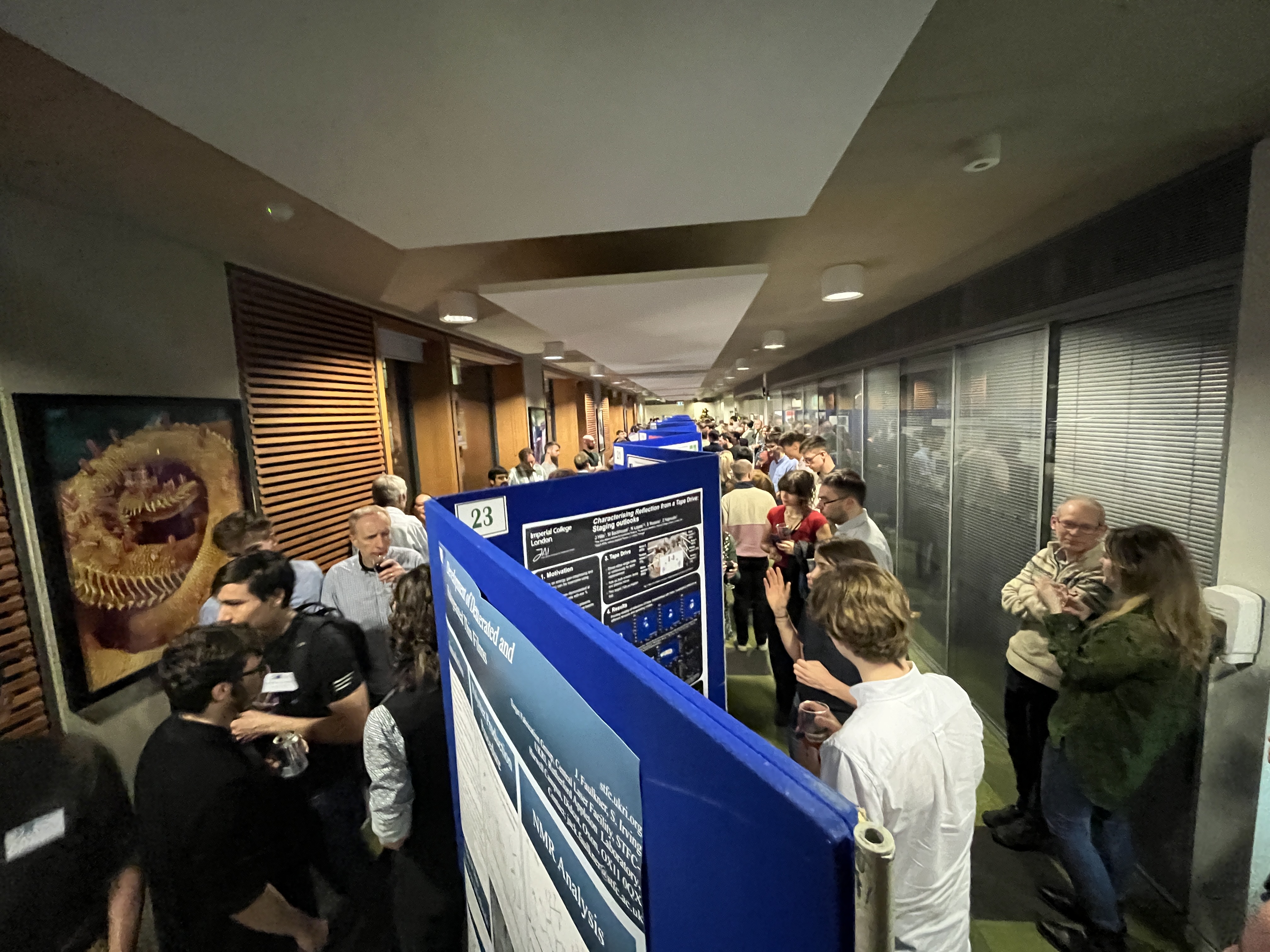 long room with blue zig-zagging poster boards running down the centre. The room is busy wiith people.